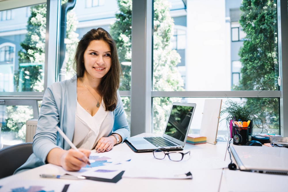 Young woman working at a desk with a laptop, drawing charts, and surrounded by office supplies, illustrating productivity and business coaching in a modern workspace.