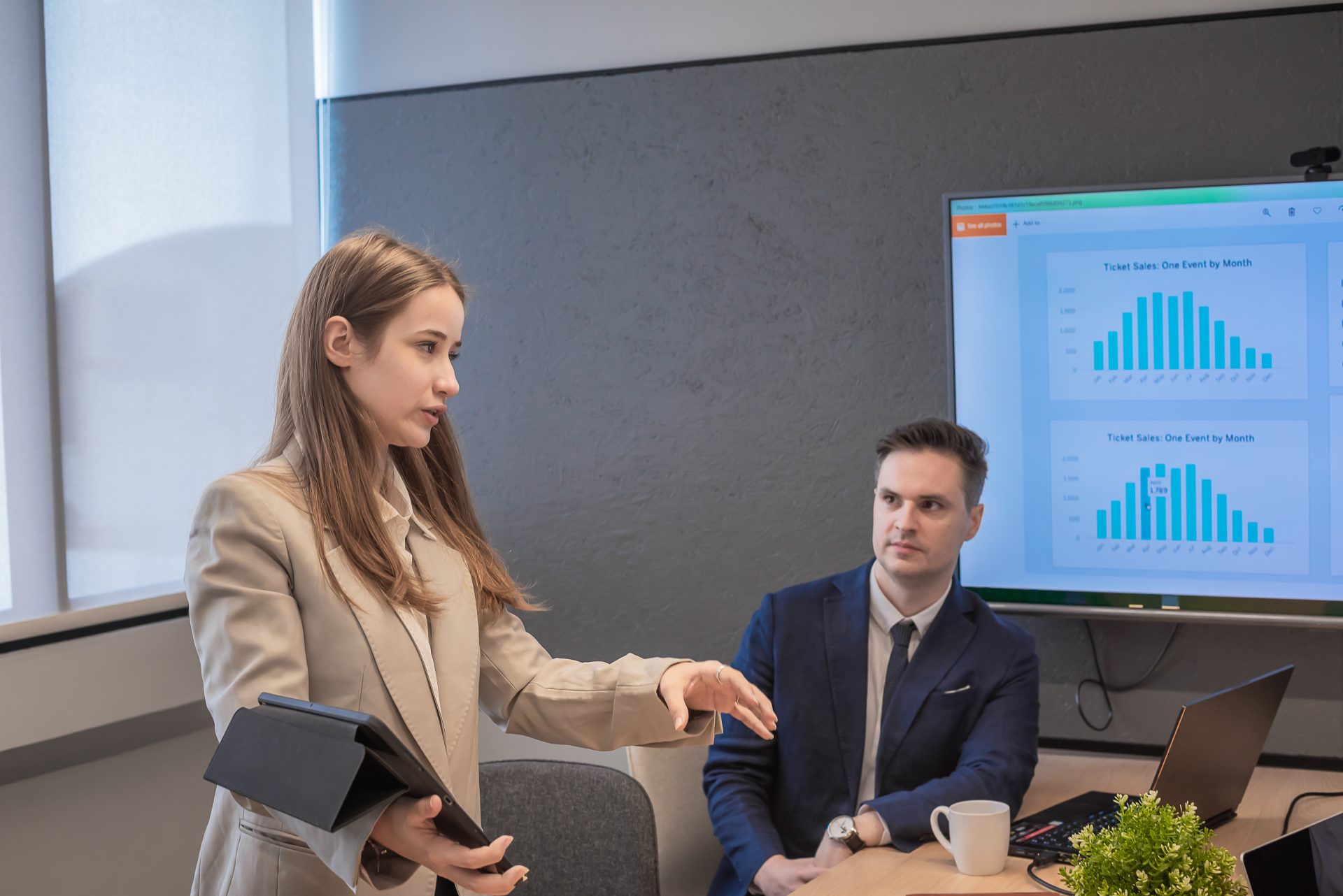 Business presentation with a woman discussing marketing strategies using a tablet, while a man observes, with charts on a screen illustrating ticket sales data.