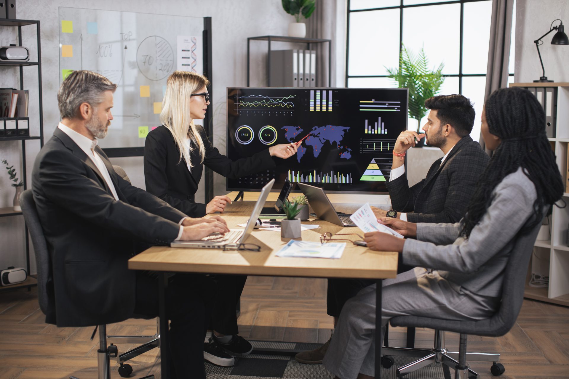 Business team meeting analyzing data presentation and strategy, featuring a diverse group engaged in discussion, with charts and graphs displayed on a screen.
